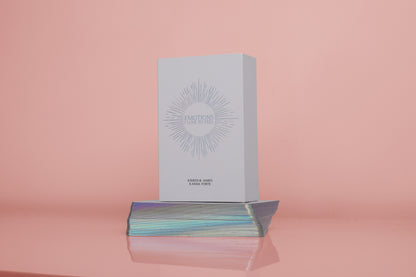 A deck of white, holographic-edged affirmation cards with a logo on the top, placed on a reflective surface with a pink background.